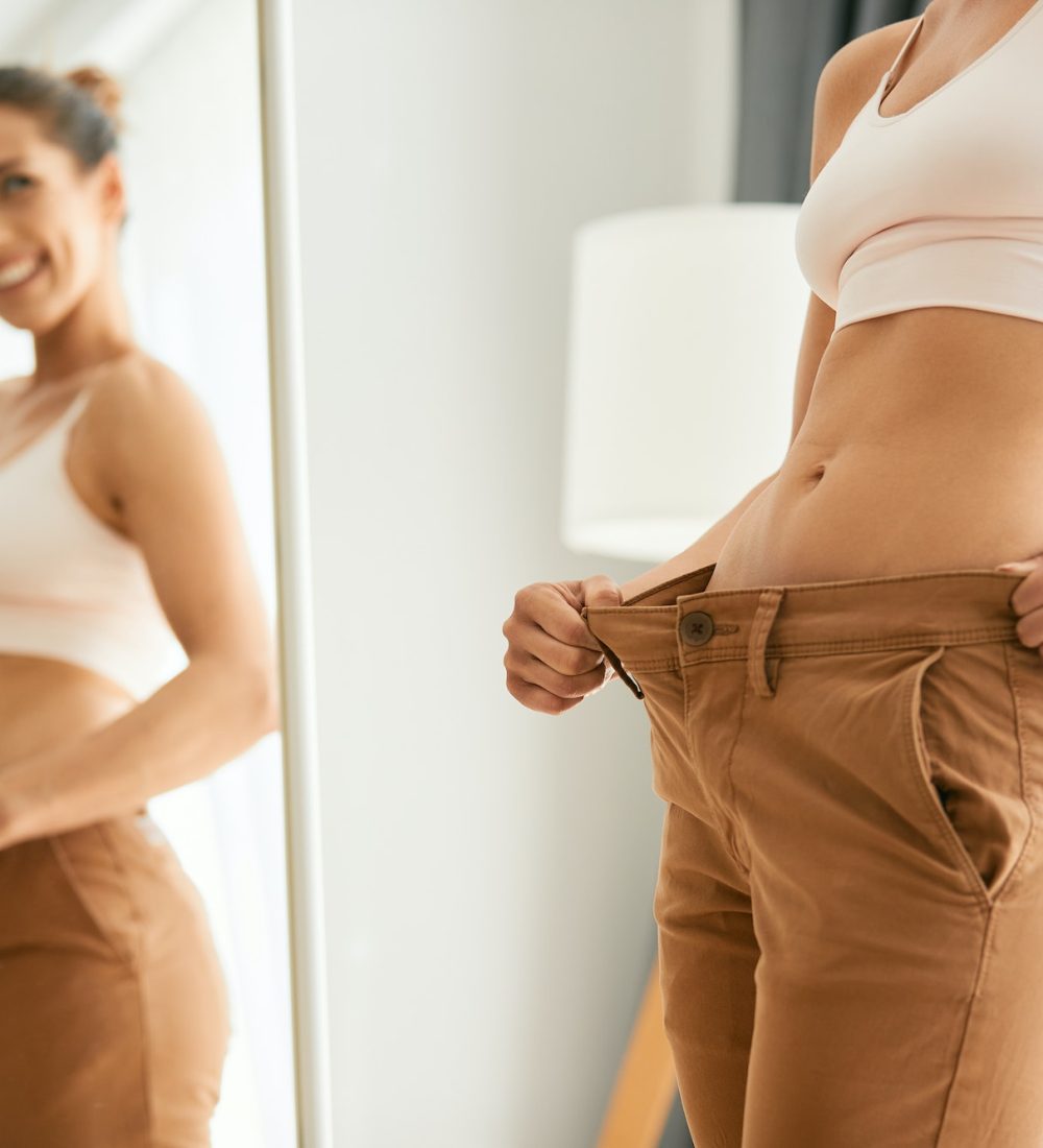 Slim woman in overweight pants looking herself in a mirror after loosing weight.